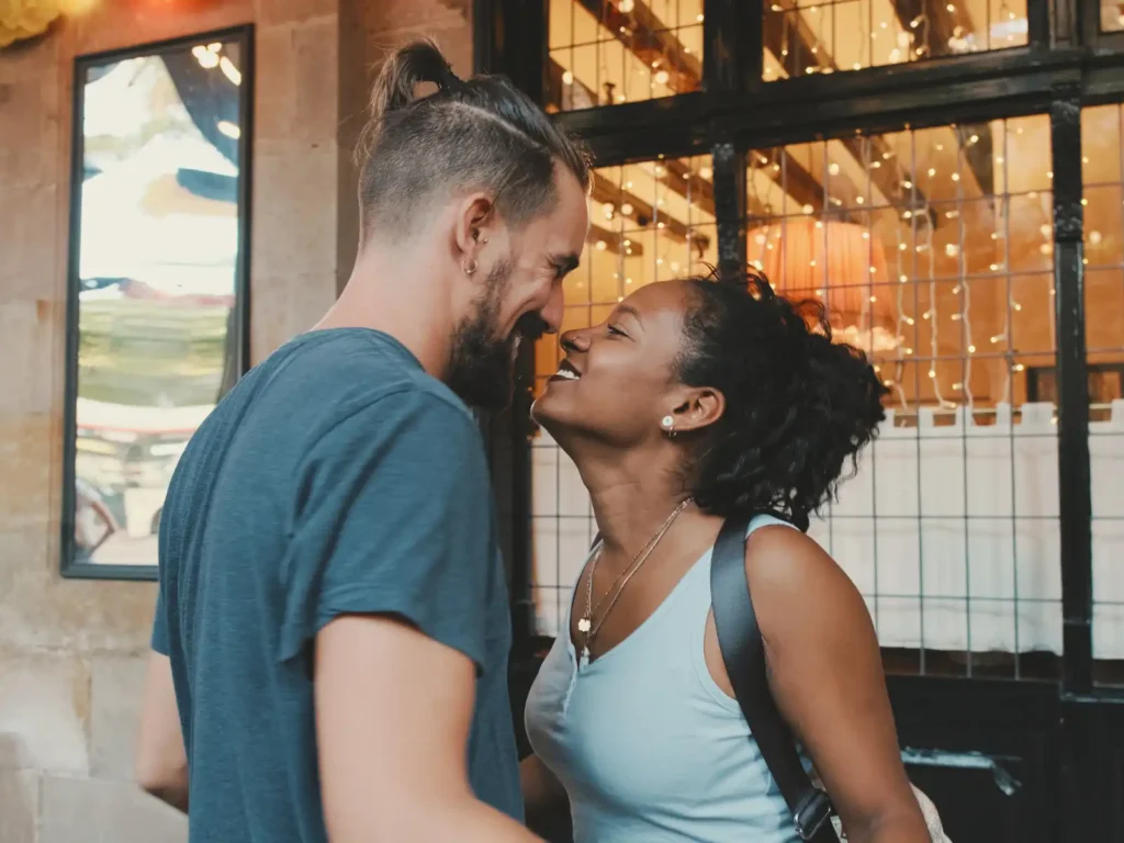 A happy couple shares a loving moment outside a warmly lit café, their faces close as they smile at each other. String lights reflect in the window behind them, creating a cozy and intimate atmosphere.