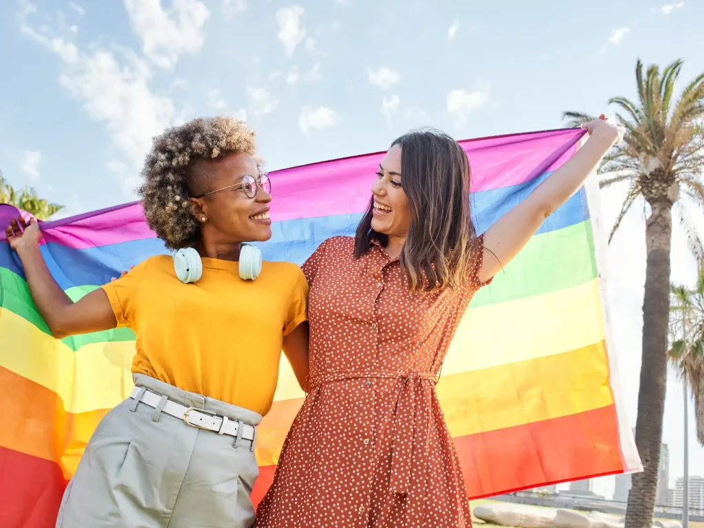 A joyful LGBTQ+ couple holding a rainbow flag at the beach in Los Angeles. LGBTQ+ couples therapy provides affirming support to strengthen relationships and navigate challenges.