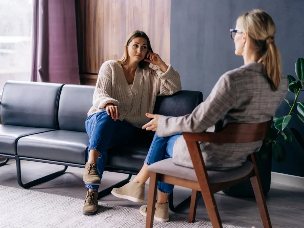 A young woman sits on a modern black leather couch, attentively listening to a professional therapist in a private consultation room, representing psychological assessment services in Los Angeles.