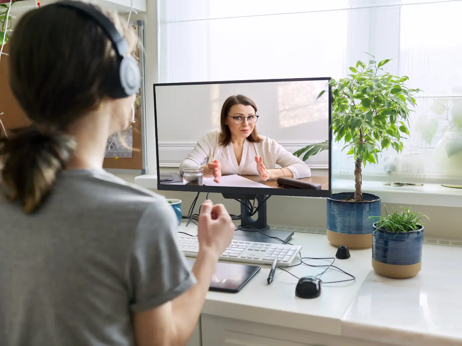 A person wearing headphones participates in an online counseling session from a home office. A therapist, visible on the computer screen, gestures as she speaks, creating a supportive and professional atmosphere. The workspace is bright, featuring natural light and green plants.